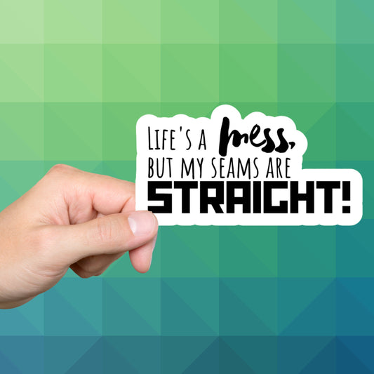 Hand holding a Life's a mess but my seams are straight sewing sticker on a green background
