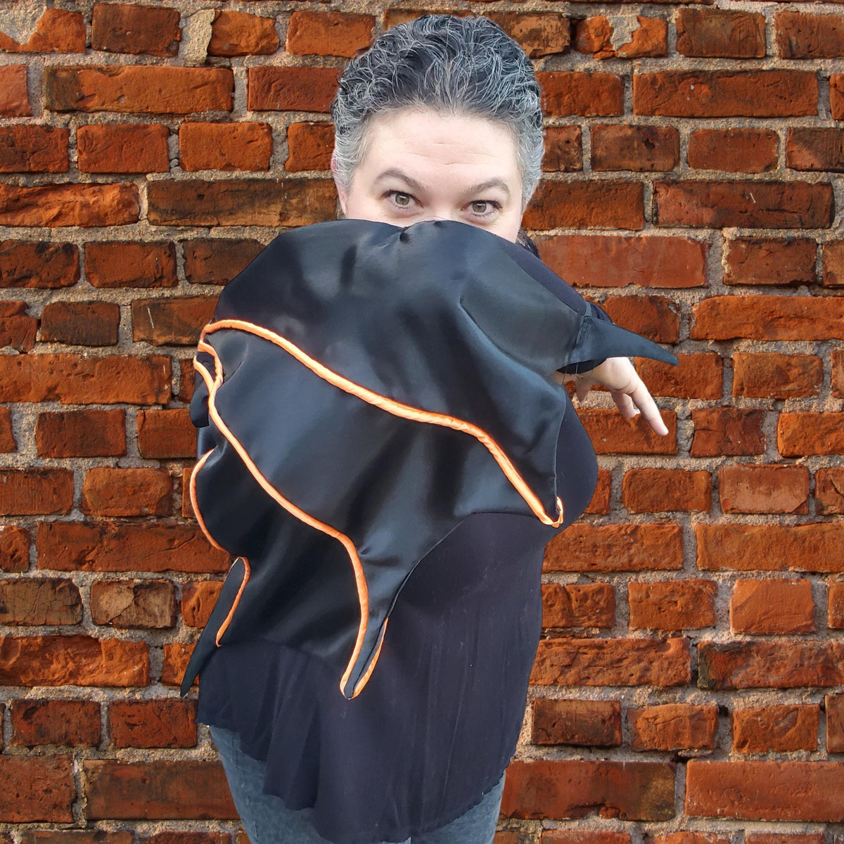 Woman holding a black bat wing cape with orange piping across her face in front of a brick wall