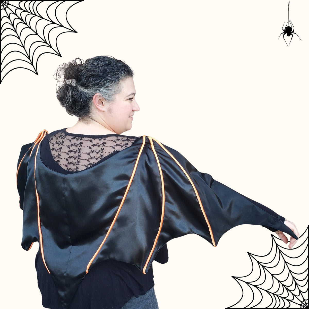 Back view of a woman with arms out showing the full wingspan of the black satin bat cape with orange piping