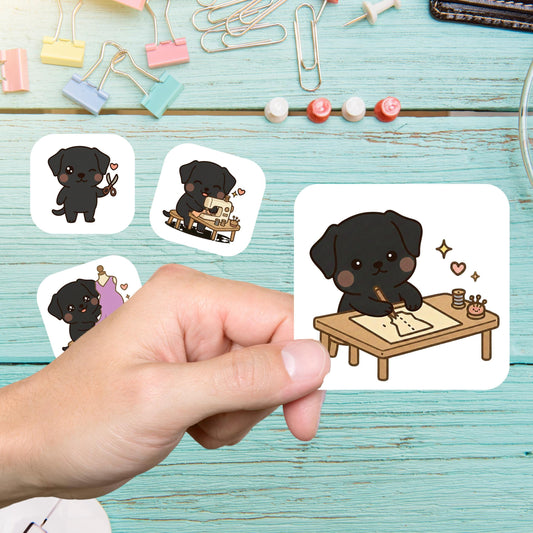 Hand holding a black lab dog sewing sticker of a dog drafting at a table, with more stickers on a teal desk