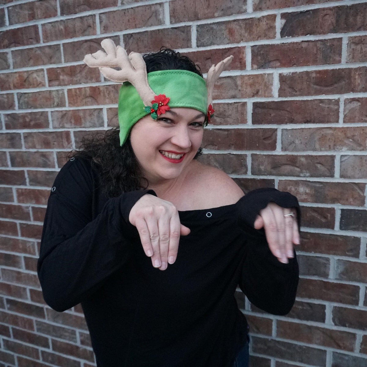 Woman wearing a green felt reindeer antler headband with holly against a brick wall