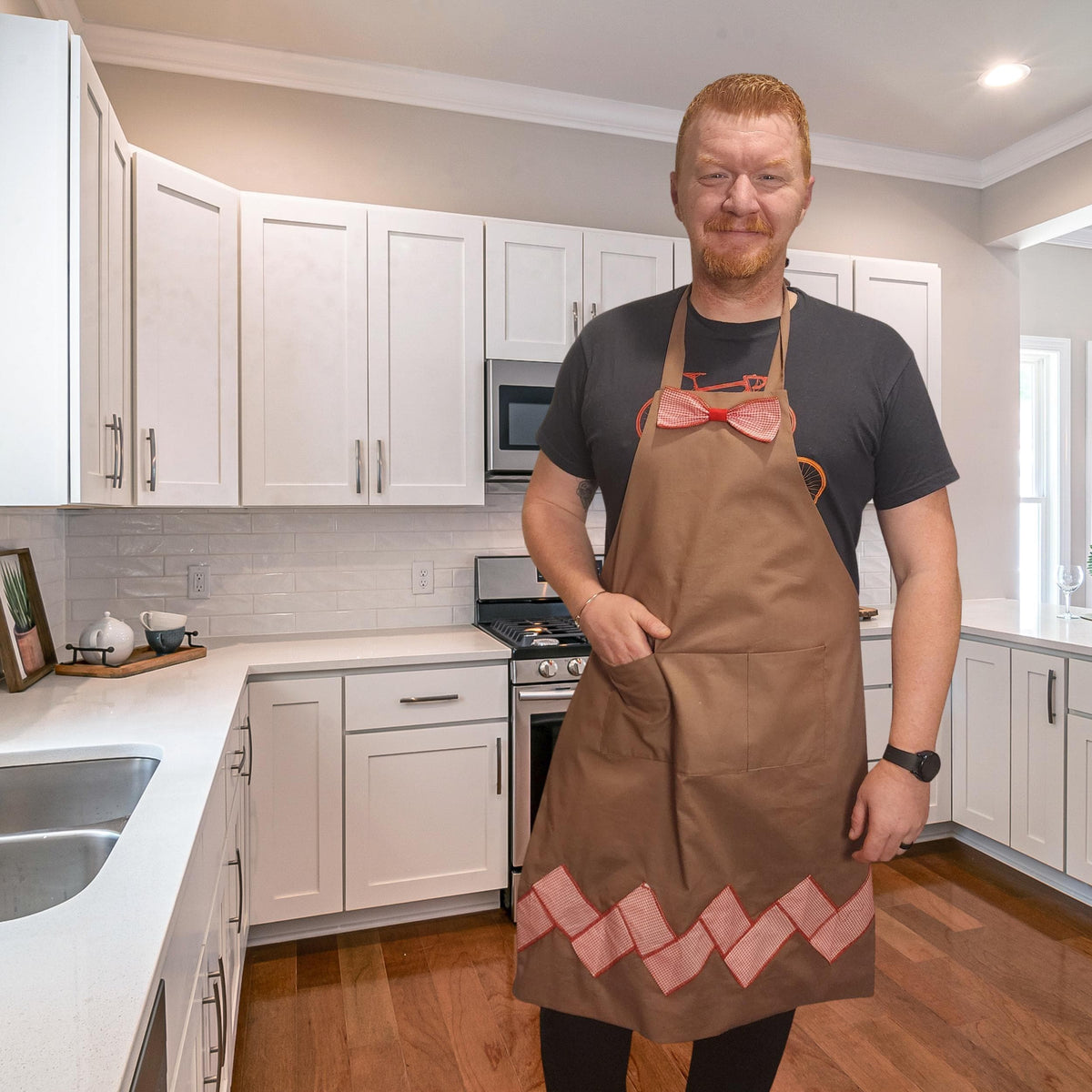 Man in a brown gingerbread apron with pink zigzag trim and bow tie in a kitchen